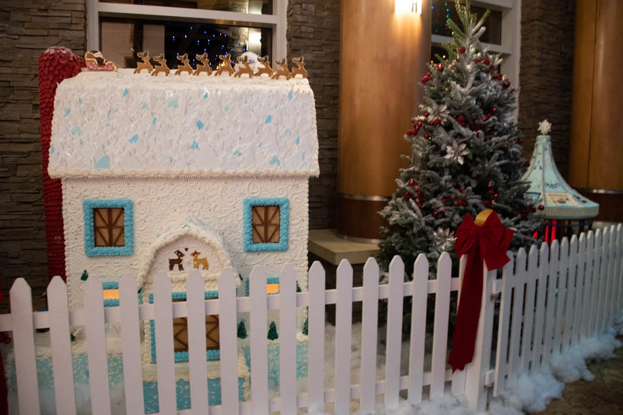 frosty white gingerbread house with blue and white icing details, part of the Turning Stone Gingerbread Village. The roof is topped with small, edible reindeer figurines lined up as if preparing for takeoff. The windows are framed in blue icing with frosted glass details, and the house sits behind a white picket fence, surrounded by fluffy white frosting “snow” and a beautifully decorated Christmas tree.