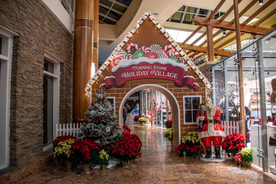 The grand gingerbread entrance to the Turning Stone Holiday Village, designed as a giant gingerbread archway decorated with candy canes, colorful peppermint swirls, and festive red ribbon accents. The words “Turning Stone Holiday Village” are prominently displayed in bold white letters on the arch. A life-sized Santa Claus figure stands beside the entrance, surrounded by bright red poinsettias, while twinkling holiday lights illuminate the display.