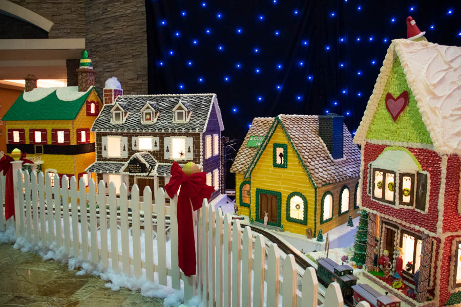 A vibrant scene showcasing a row of beautifully crafted Turning Stone gingerbread houses, each uniquely decorated with colorful frosting, candy accents, and glowing windows. The houses sit behind a white picket fence dusted with fluffy snow, and the background features a starry night sky of twinkling blue lights. A miniature train track winds through the village, adding a whimsical holiday touch to this enchanting display.