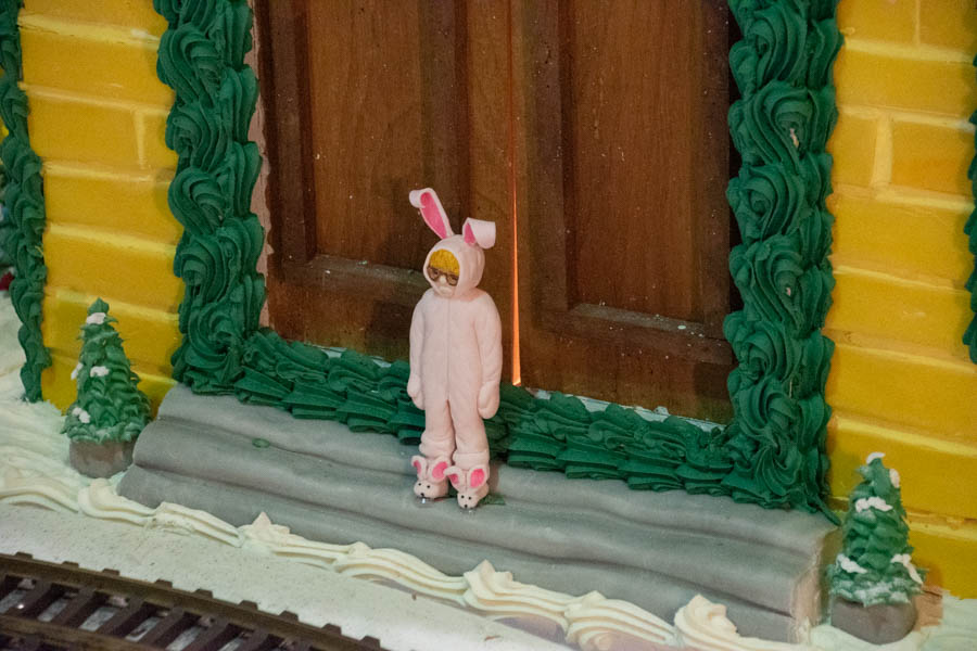 A close-up of a small figurine dressed in a pink bunny suit standing in front of the doors of a yellow gingerbread house. The house is decorated with green piped icing trim around the windows and doors, with snowy frosting on the steps, adding a charming touch to this holiday scene.