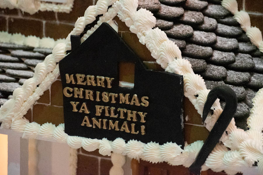 A gingerbread house porch featuring a black fondant sign that reads, “Merry Christmas Ya Filthy Animal!” in bold gold lettering. The sign is surrounded by frosted icing “snow” and candy cane decorations, paying homage to the classic line from Home Alone.