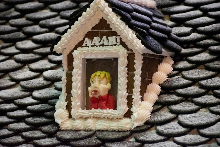 A playful detail on a gingerbread house roof featuring a fondant figure of a blonde boy with his hands on his cheeks, shouting “AAAH!” as piped letters spell out the famous Home Alone scream scene. The roof is carefully decorated with black frosting shingles dusted with powdered sugar snow.