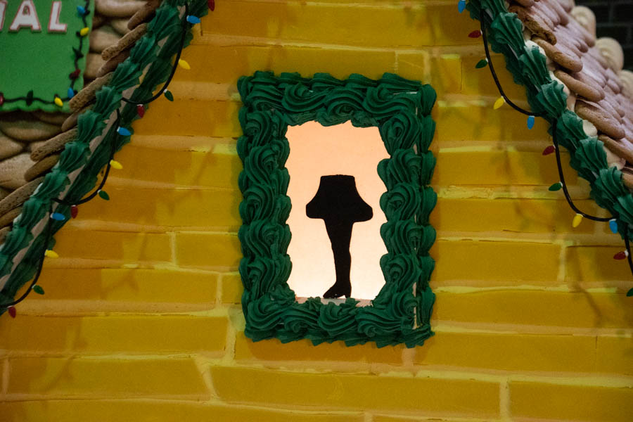 A yellow gingerbread house with a window showing a silhouette of the iconic leg lamp from A Christmas Story. The window is framed in green icing and surrounded by festive holiday lights piped onto the house exterior, adding to the nostalgic charm.