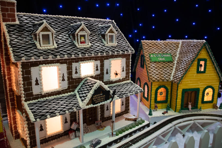 A large, two-story gingerbread house with intricate details, including a snowy white frosting roof made of carefully piped icing shingles. The house features lit-up windows adorned with festive silhouettes like Christmas trees and holiday decorations. A small porch with candy cane columns welcomes visitors, and a miniature train track loops around the house, adding a whimsical touch to the winter scene