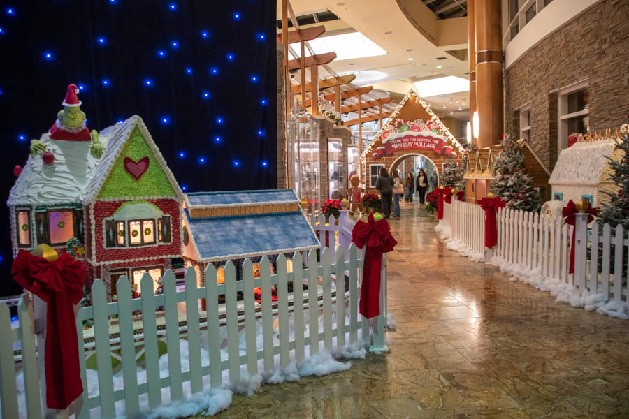 A wide shot of the Turning Stone Gingerbread Village, featuring colorful gingerbread houses behind a white picket fence. The scene includes houses with red bows, snowy frosting details, and twinkling blue lights in the background. A red and green Grinch-themed house stands prominently with festive decorations.