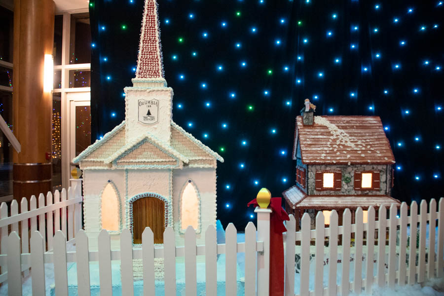 A festive scene featuring the Turning Stone gingerbread chapel with a tall, red-spired roof and glowing windows, surrounded by snowy frosting. Next to the chapel is a stone-textured gingerbread house with envelopes scattered across the roof and a small figure at the chimney holding a list.