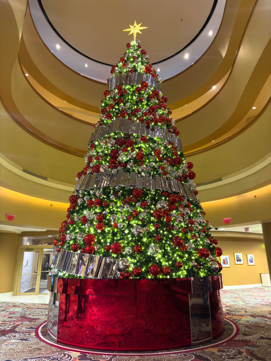 A massive Christmas tree at Turning Stone Resort, glowing with twinkling white lights, red and silver ornaments, and a golden star perched at the very top. The tree is surrounded by reflective silver panels and a vibrant red base, perfectly complementing the opulent holiday décor.