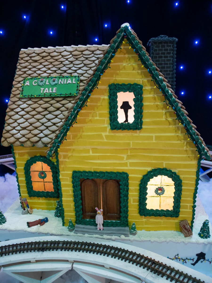 A bright yellow gingerbread house at Turning Stone designed to resemble a classic colonial-style home. The frosted shingle roof has snowy dusting, and green icing decorates the edges like holiday garlands. The lit windows feature a festive silhouette of a leg lamp, paying homage to A Christmas Story. Small holiday figurines and a tiny nutcracker are placed outside the entrance, surrounded by snowy frosting, completing the charming gingerbread display