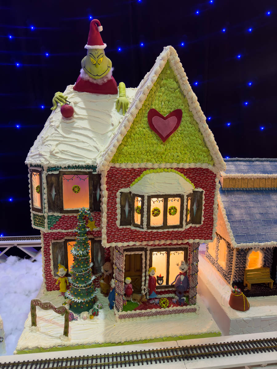 A whimsical Grinch-themed gingerbread house with green and red icing. The Grinch himself sits on the snowy roof, wearing a Santa hat and clutching a red ornament. The house features glowing windows, a heart decoration on the façade, and a detailed frosting Christmas tree outside with fondant figurines.