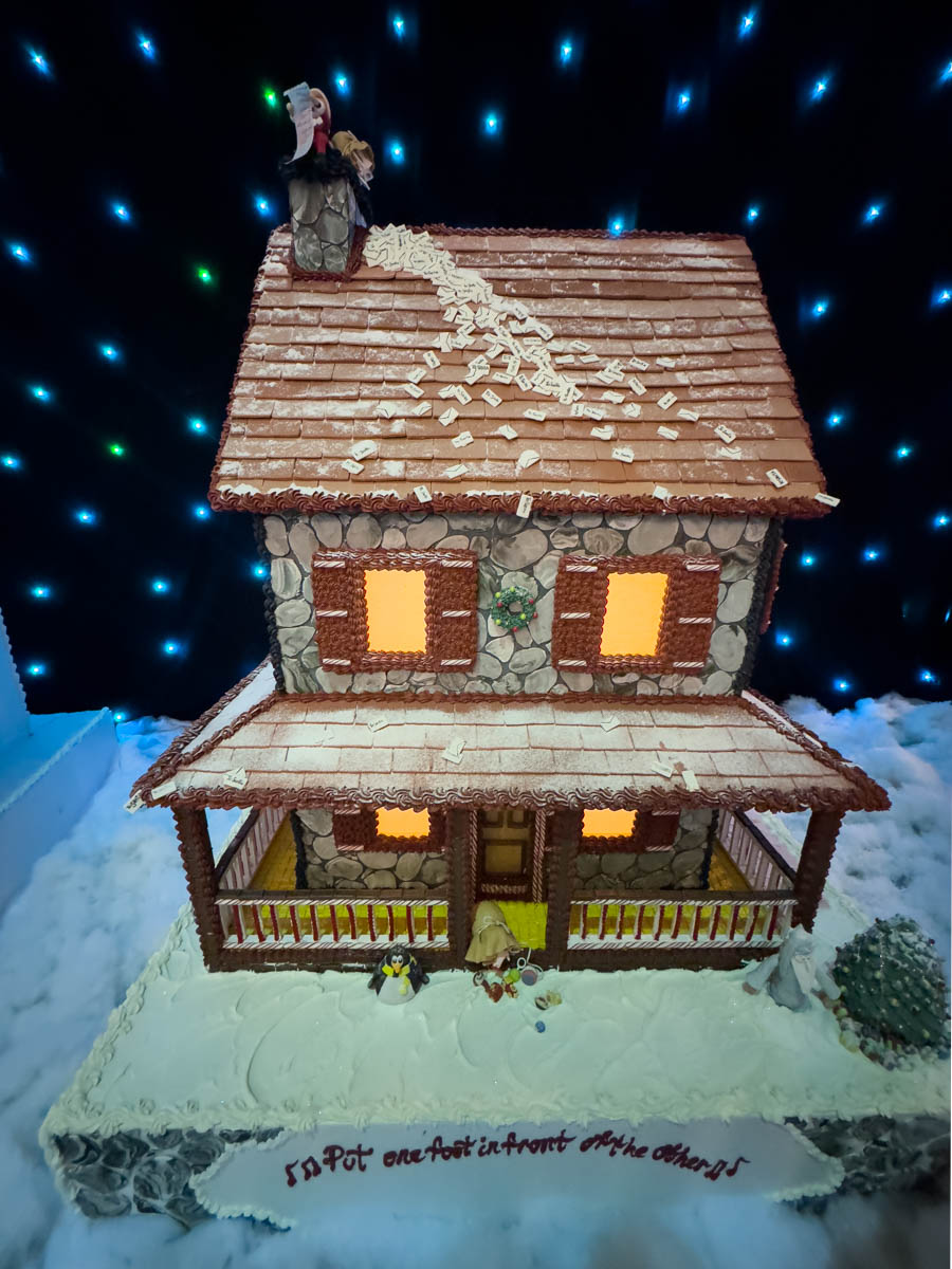 A stone-textured Turning Stone gingerbread house with glowing orange windows that bring warmth to the frosty setting. The house features realistic details, including piped stone walls and a roof covered with brown icing shingles dusted with powdered sugar snow. A whimsical figure appears at the top of the chimney, holding a long list of names, and tiny penguin figurines stand on the snowy frosting base near the porch, adding a playful element.