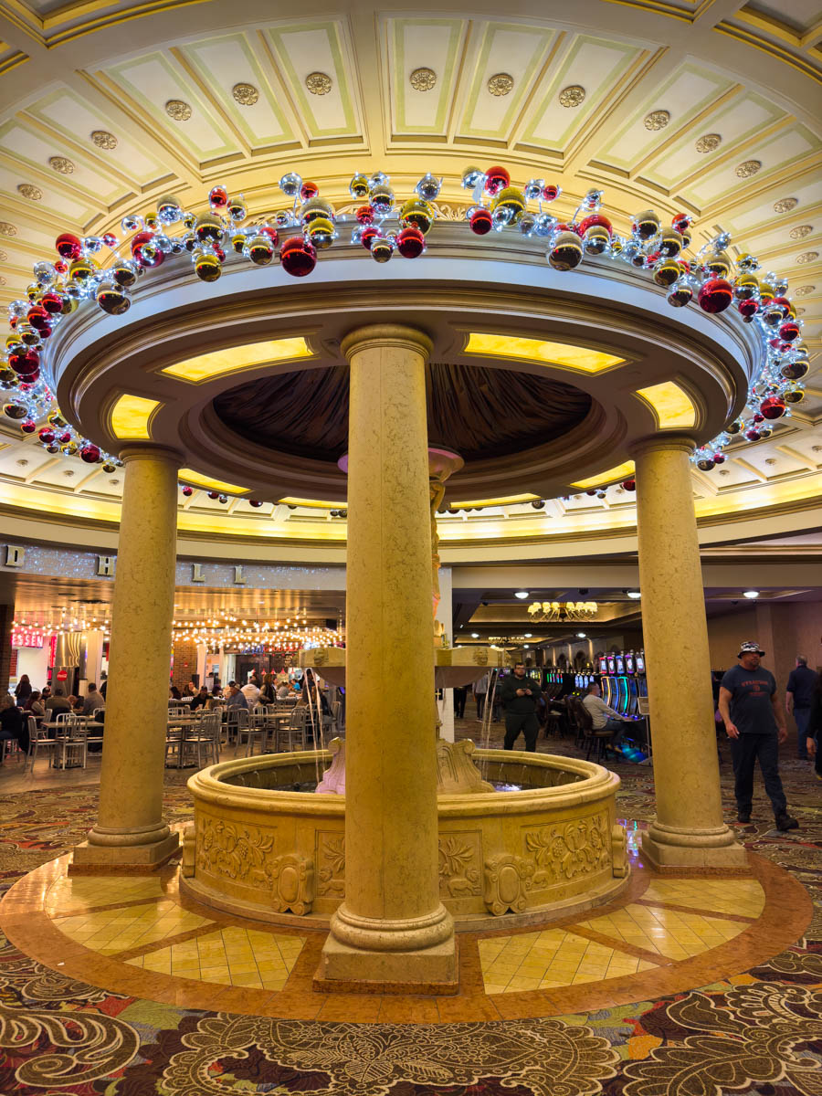 A stunning circular fountain display inside Turning Stone Resort, framed by large marble columns and topped with intricate molding. Above the fountain hangs a festive arrangement of oversized red, gold, and silver Christmas ornaments, creating an elegant and luxurious holiday atmosphere. Visitors are seen enjoying the space in the background.