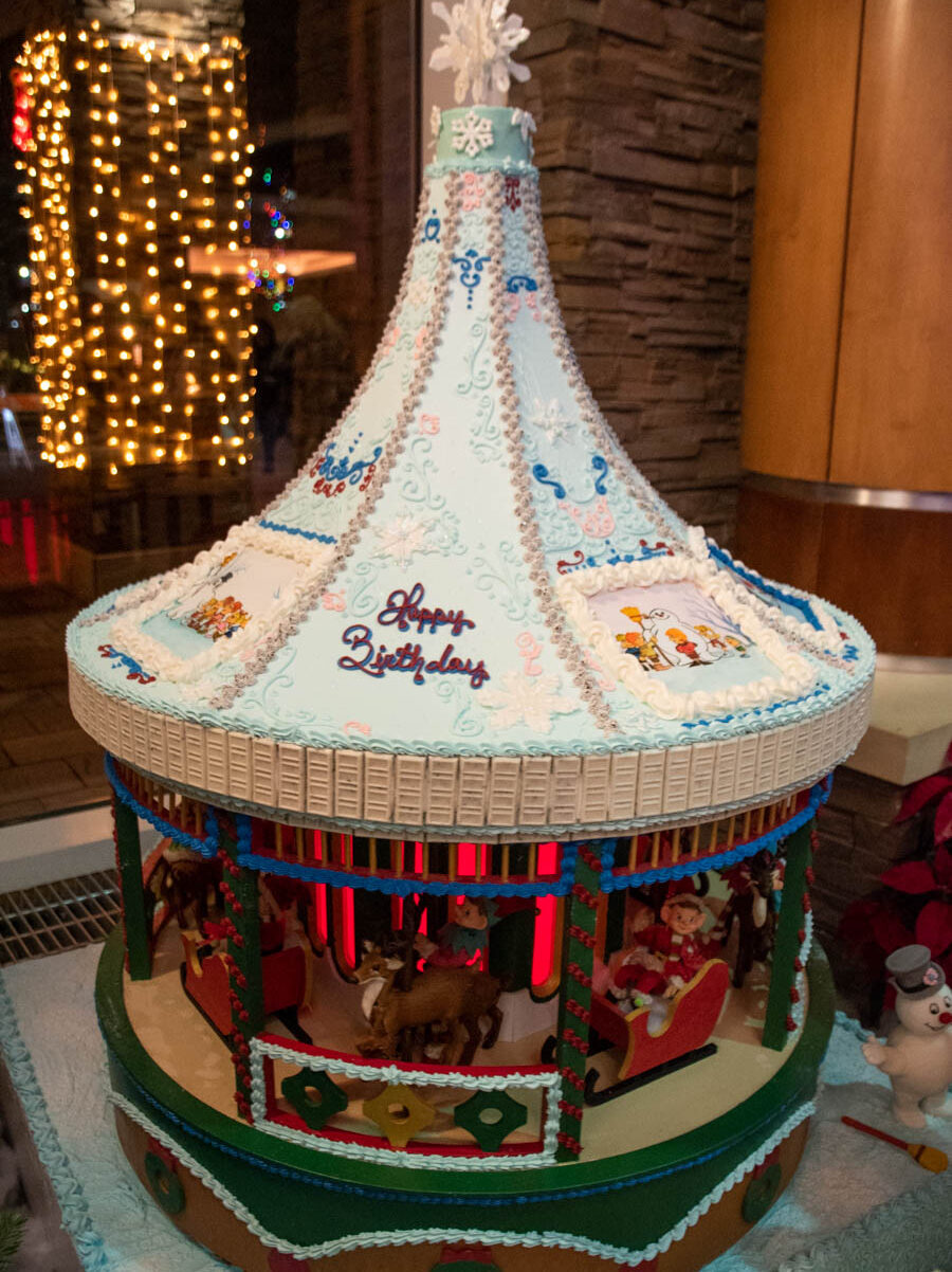 n elaborately decorated gingerbread carousel at the Turning Stone Holiday Village with a detailed, towering blue and white frosting roof, adorned with intricate snowflakes, swirls, and festive designs. The words “Happy Birthday” are piped in red icing. Beneath the roof, a carousel of holiday-themed figurines includes reindeer, elves, and Santa riding in a sleigh. Red glowing lights inside the carousel illuminate the figures, adding a magical and festive atmosphere.