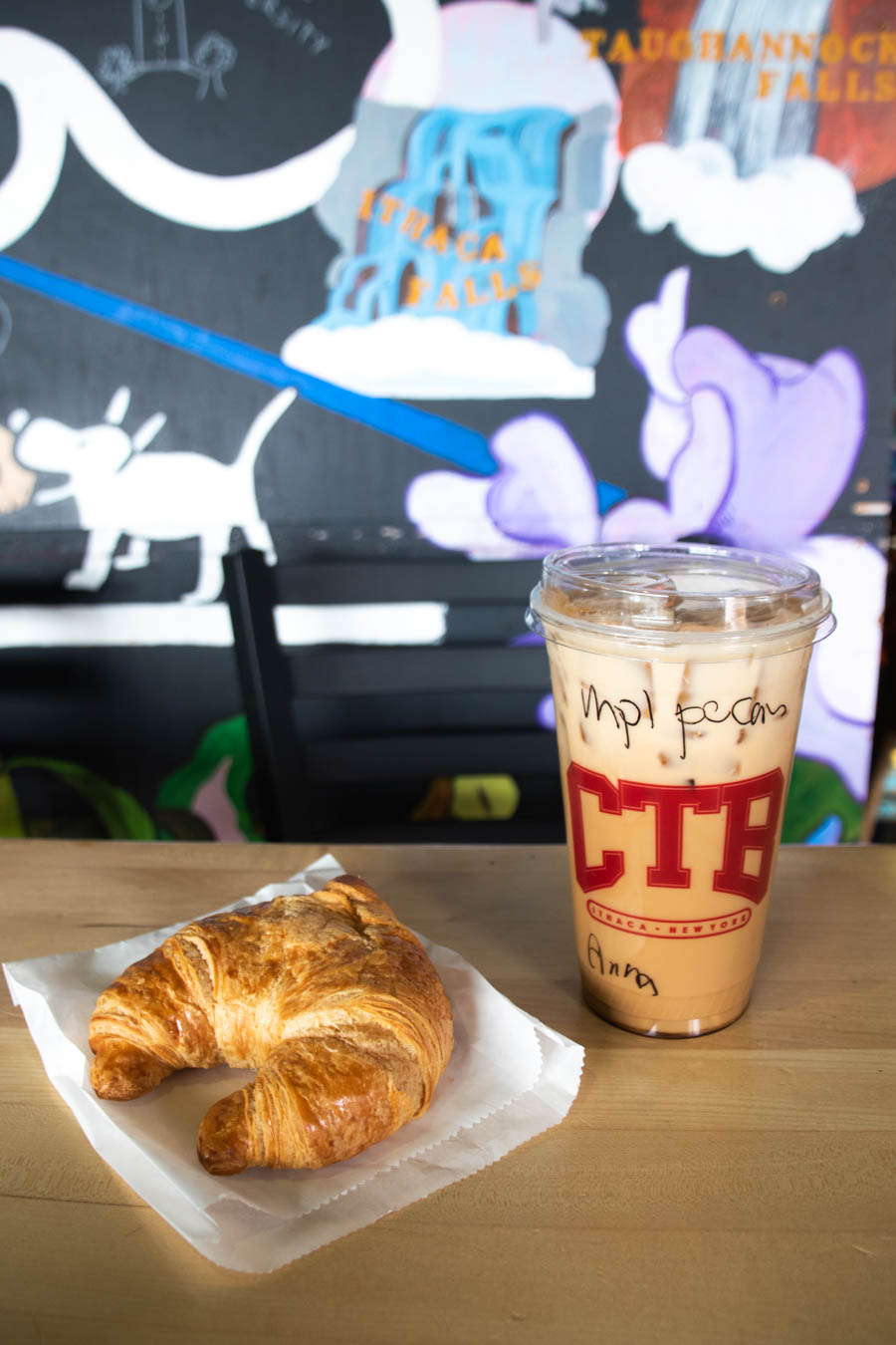 Image of a croissant and iced coffee with "CTB" logo – A flaky croissant and a maple pecan iced coffee from a local café, set against a colorful mural featuring Ithaca Falls, a great stop during a chilly adventure.