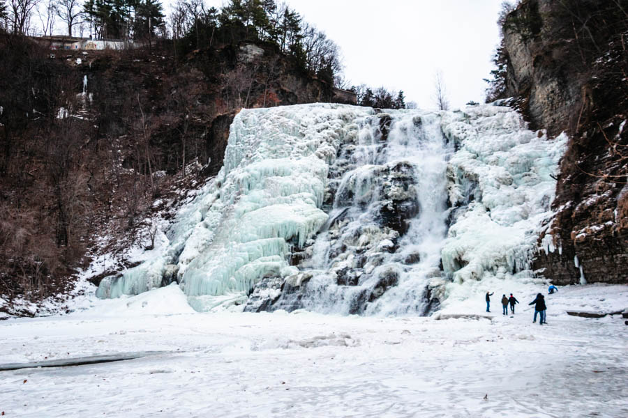 Image of a frozen Ithaca Falls with people standing nearby – A breathtaking frozen waterfall in the Finger Lakes, with visitors admiring the icy cascade surrounded by snow-covered cliffs.