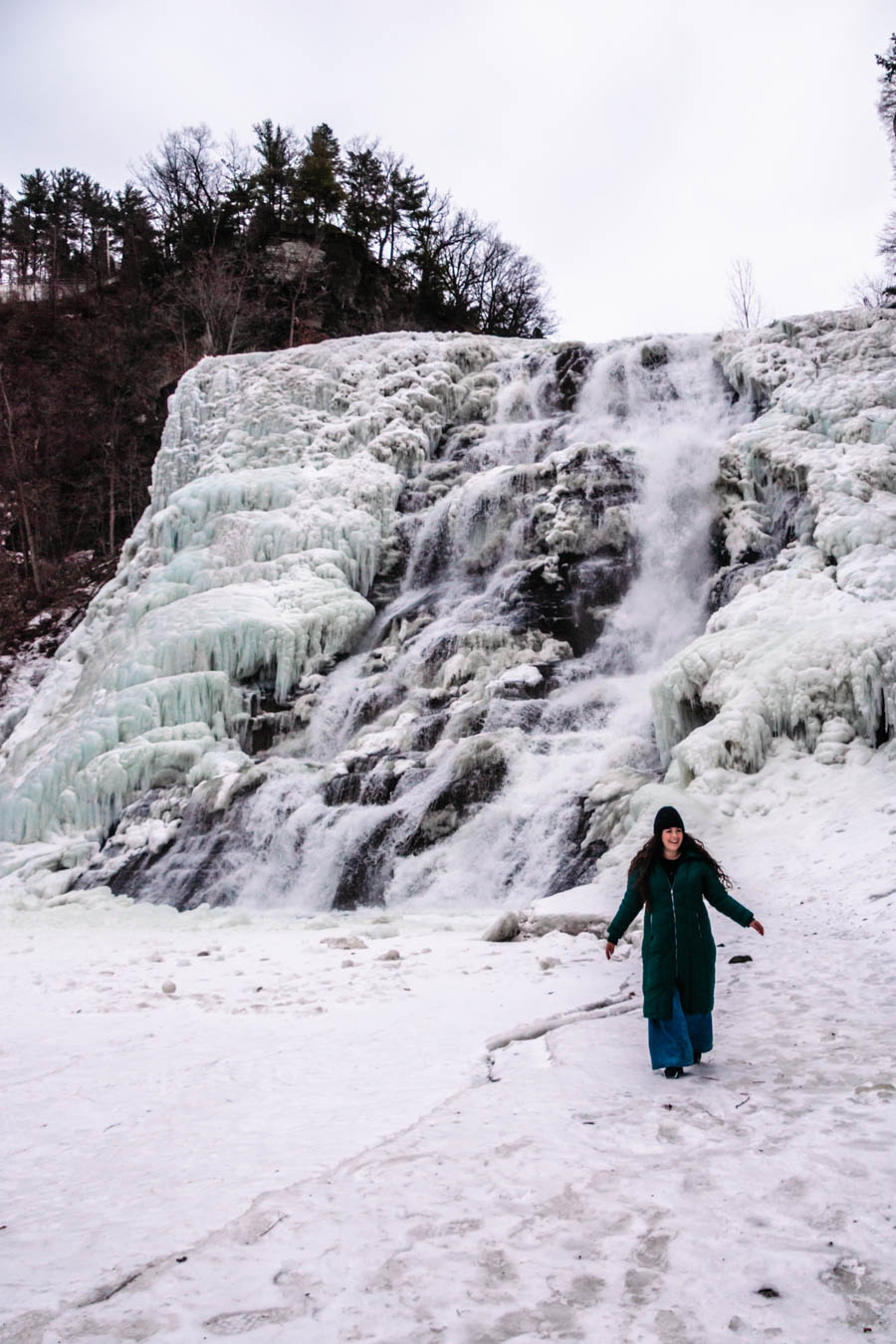 Image of a woman walking across the snowy ground near a frozen waterfall – A winter adventurer takes in the magic of a frozen waterfall, walking carefully across the snowy landscape with towering ice formations in the background.