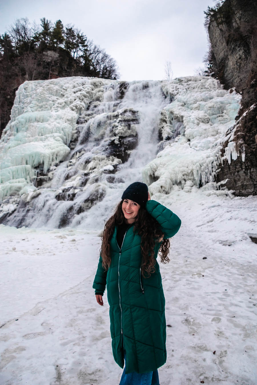 Image of a woman standing close to a partially frozen waterfall – A traveler in a green winter coat enjoys the stunning view of a cascading frozen waterfall, surrounded by glistening ice formations.