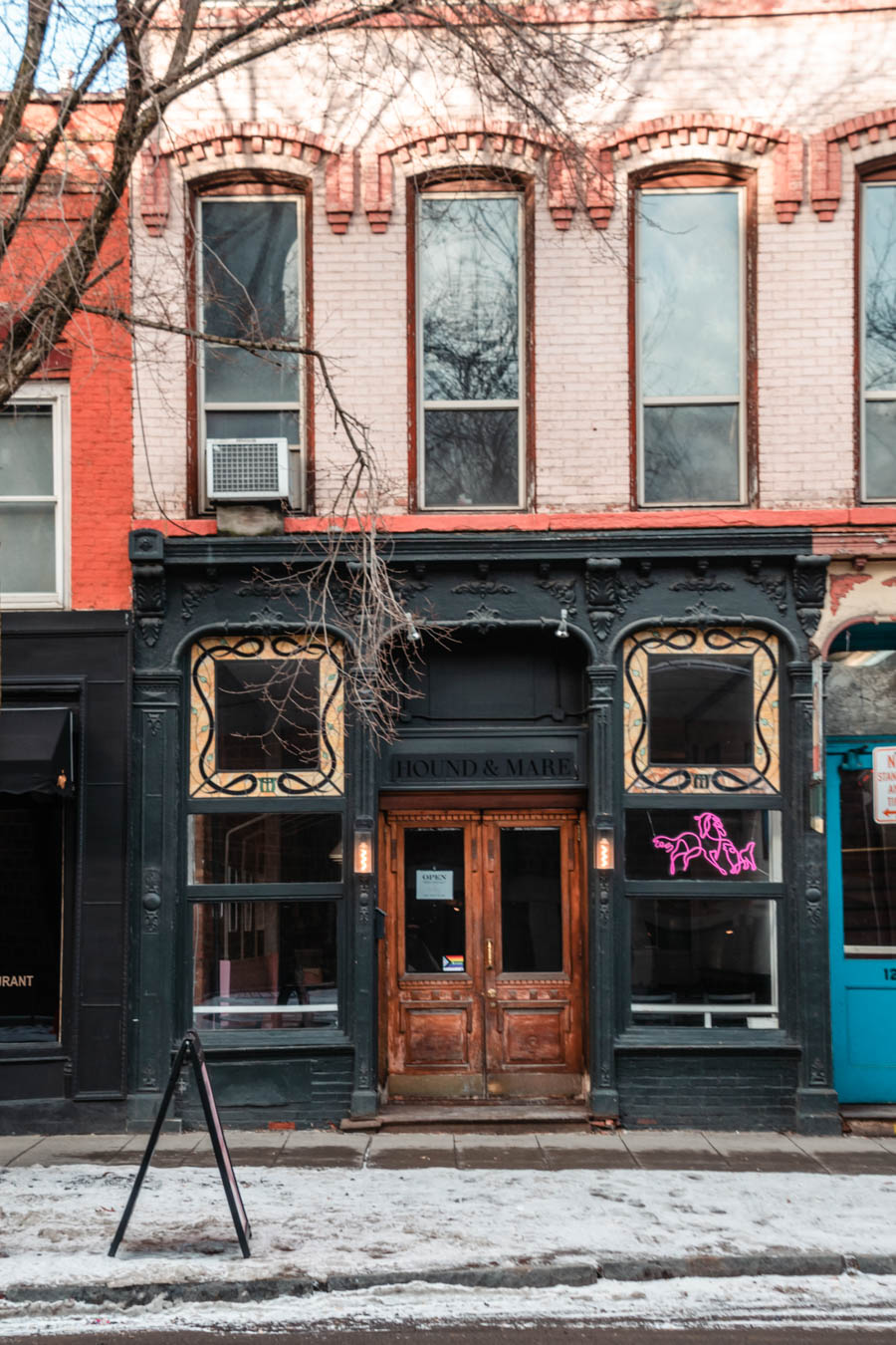 Image of a rustic storefront with a neon dog sign reading "Hound & Mare" – The charming black-and-gold facade of Hound & Mare, a cozy café in downtown Ithaca, with its neon dog logo glowing on a winter day.