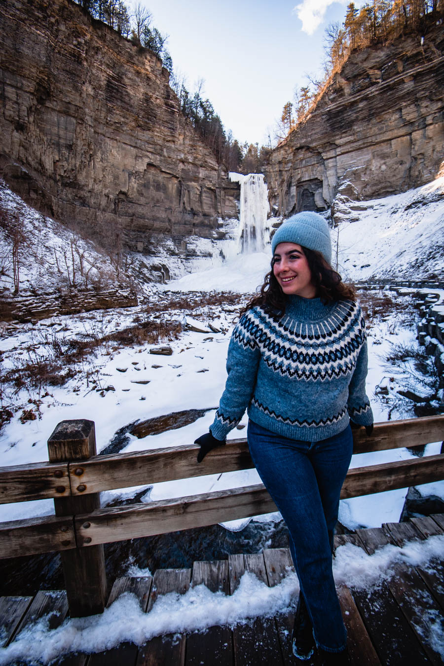 Image of a woman in a blue knit sweater and beanie, smiling in front of a frozen waterfall – A traveler dressed in cozy winter wear smiles on a wooden bridge with a stunning view of Taughannock Falls in the background.