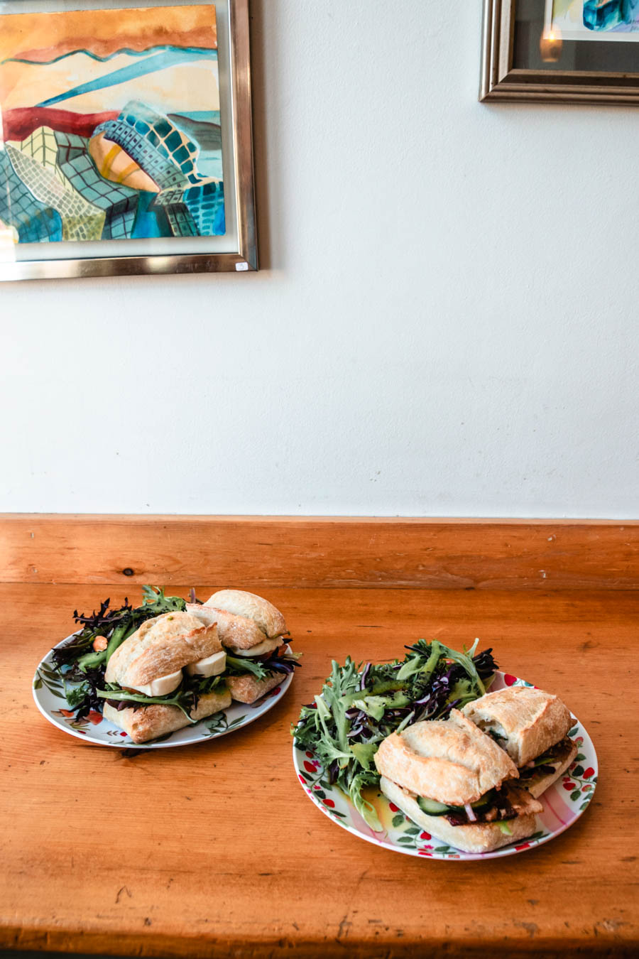 Image of two plates with sandwiches and salads on a wooden counter – Fresh sandwiches and mixed green salads served on floral plates in a cozy café, with colorful artwork hanging on the wall.