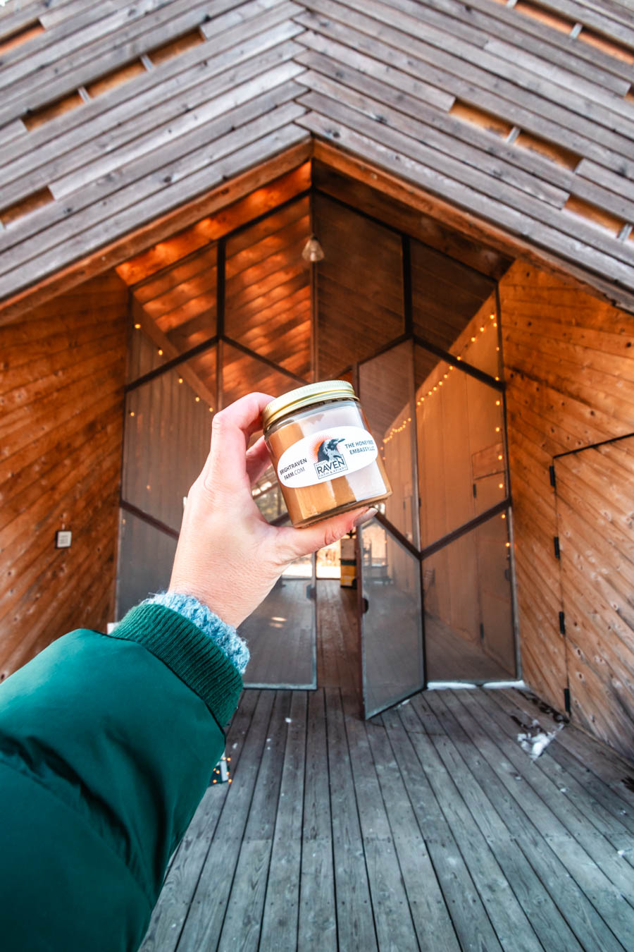 Image of a hand holding a jar of honey in front of a modern wooden building with geometric glass doors – A jar of locally produced honey from a Finger Lakes farm shop, with a stunning wooden and glass storefront in the background.