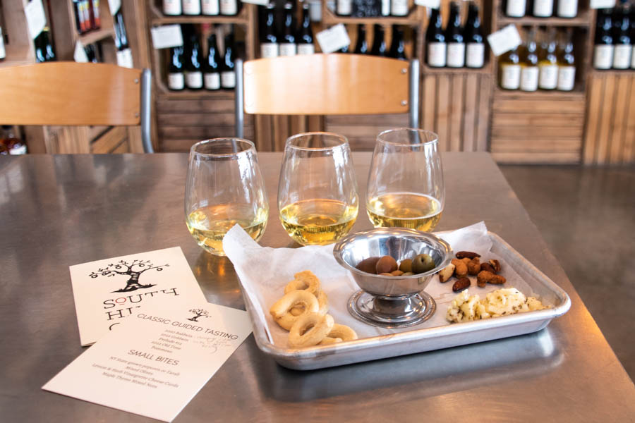 Image of a cider tasting setup with small bites at South Hill Cider – A cider tasting experience in the Finger Lakes featuring three glasses of white wine, olives, cheese, and snacks, creating the perfect winter indulgence.