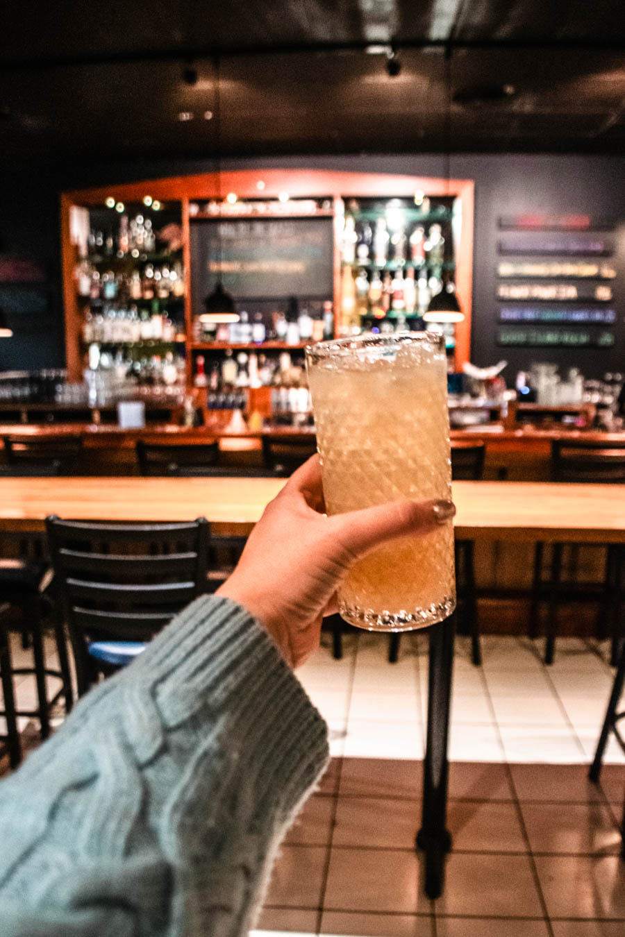 Raising a toast at a Finger Lakes bar – A hand holding a textured glass of a golden cocktail in a warmly lit bar.