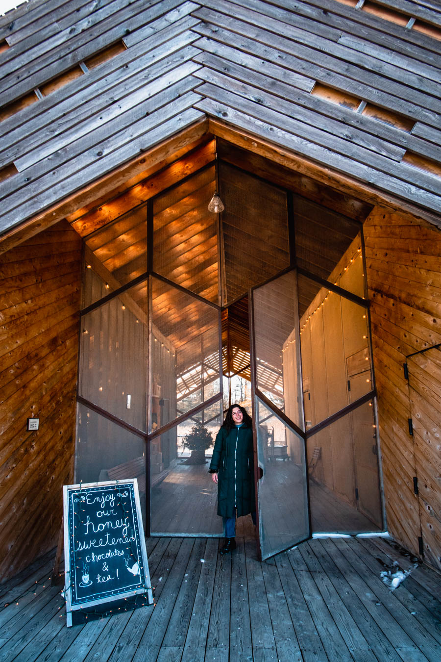 Image of a woman walking toward the entrance of a honey and tea shop with twinkling string lights – A winter visitor stepping into a beautifully designed wooden farm shop, welcomed by a sign inviting guests to enjoy honey-sweetened treats.