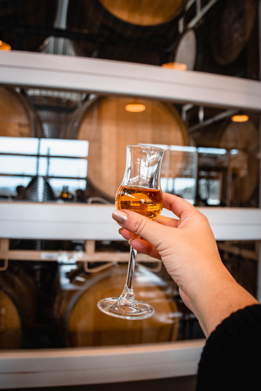 Image of a hand holding a glass of amber-colored liquor in front of wooden barrels – A close-up of a hand holding a glass of aged spirit, with a background of oak barrels at a Finger Lakes cidery.