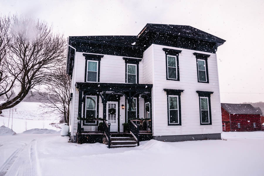 A charming snow-covered farmhouse with black trim, capturing the picturesque countryside of the Finger Lakes in winter. 