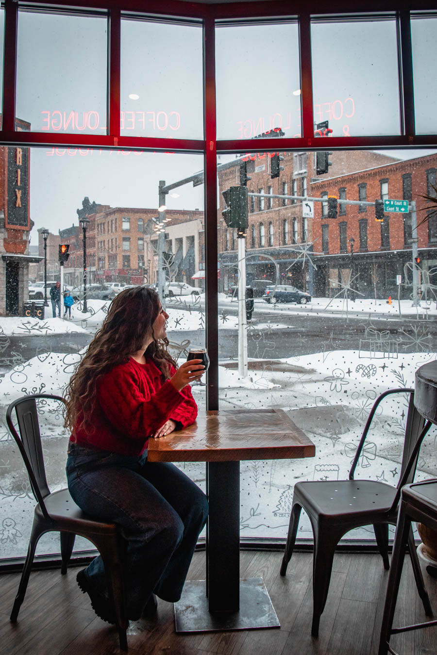 A woman in a red sweater sipping beer inside the warm BRU64, looking out at the snowy streets of Cortland, NY through large windows.