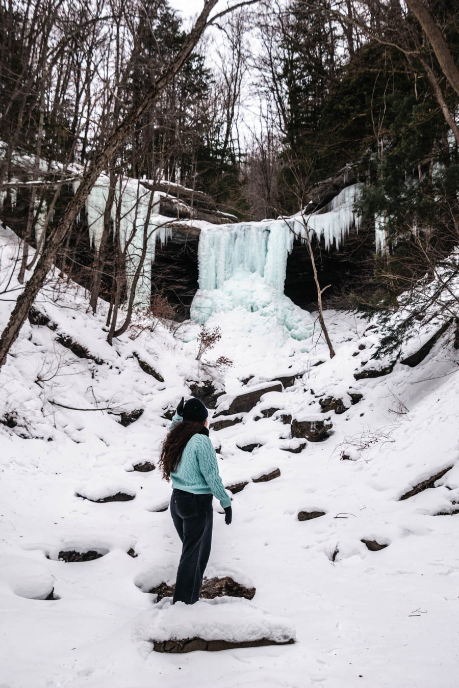 Woman standing in front of a breathtaking view of a frozen waterfall framed by snow-covered trees, showcasing the beauty of the Finger Lakes in winter.