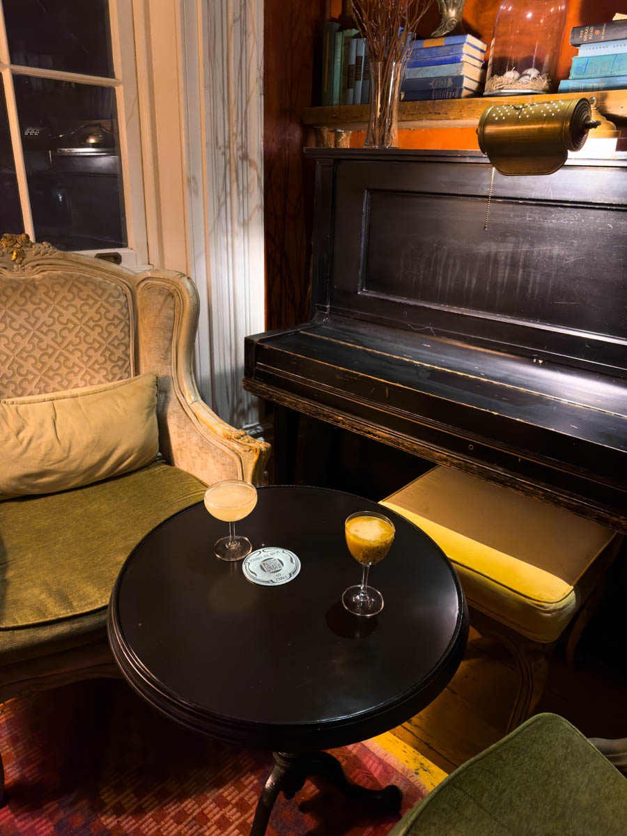 Vintage-style bar seating with cocktails (Bar Argos) – A cozy lounge with a vintage piano, plush armchairs, and two elegant cocktails on a black table.