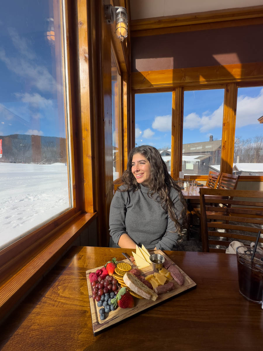 Enjoying a charcuterie board with a scenic mountain view – A woman sits by a large window with a beautifully arranged charcuterie board, enjoying the breathtaking winter scenery.